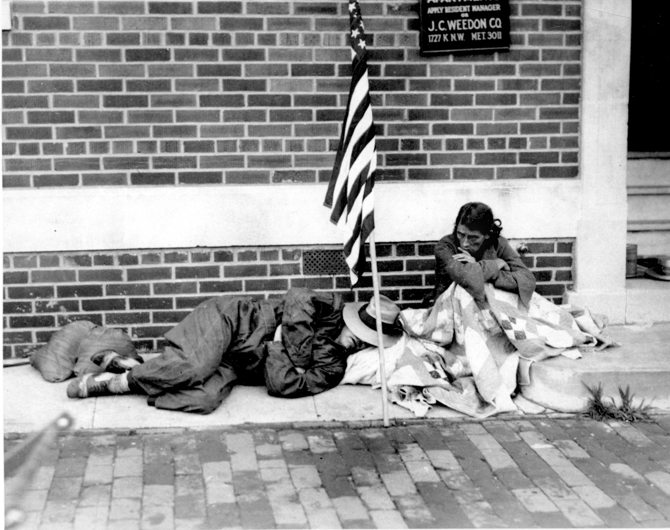 A war veteran sleeps on the sidewalk as his wife sits wrapped in blankets in Washington D.C. on July 29, 1932. Photo | AP