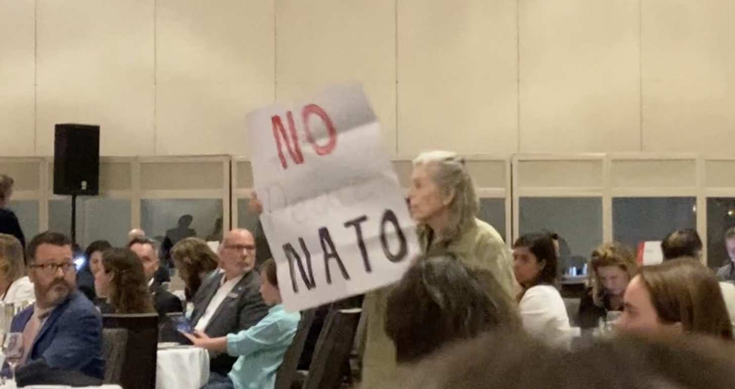 Montreal activist Laurel Thompson (a woman with grey hair and a jacket) holds up a NO NATO sign facing the stage where public-relations presentation is taking place.