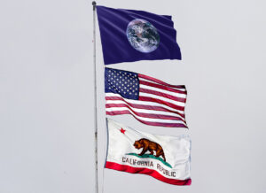 A flag pole with the Earth flag flying on top depicting the “Blue Marble” image of the Earth, photographed from the Apollo 17 spacecraft, in 1972. The American flag flies underneath that on the flag pole and the California state flag under that.