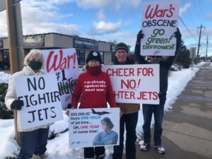 Activiști stau în fața biroului parlamentarului Terry Dowdall ținând pancarte care protestează împotriva achiziționării planificate de avioane de luptă de către Canada