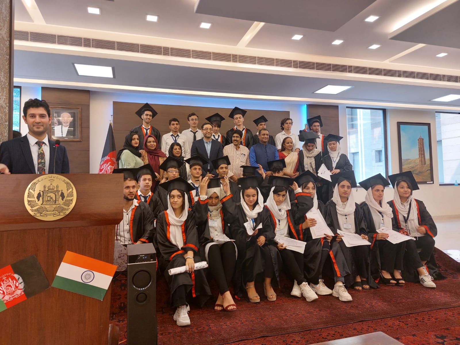 World BEYOND War Afghanistan chapter coordinator Dr. Nazir Ahmad Yosufi stands at a podium at left, with rows of students sitting at right in their graduation caps and gowns holding their diplomas.