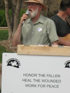 アッパー中西部支部コーディネーターのフィル・アンダーソン氏がマイクに向かって話します。 正面には「戦没者を讃え、負傷者を癒し、平和のために働け」と書かれた「平和のための退役軍人会」の看板がある。
