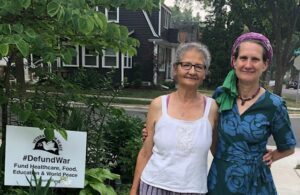 World BEYOND War Koordinatorke Madison Chapter Stefania Sani i Janet Parker poziraju za fotografiju na ulici sa drvoredima sa natpisom #DefundWar Veterans For Peace u pozadini.