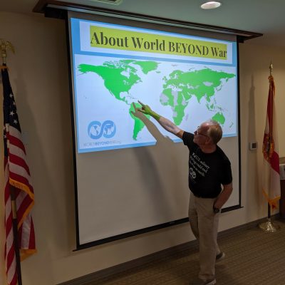 Al Mytty points to a projector screen where a map of the world is shown with the heading "About World BEYOND War"