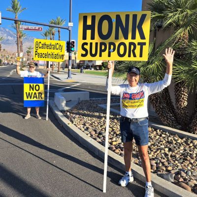 Two protesters stand at the street corner holding big yellow signs saying "Cathedral City Peace Initiative", "HONK SUPPORT", and "NO WAR" with palm trees and mountains in the background.