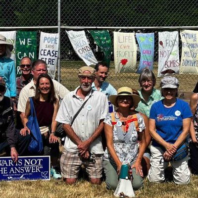 Participants from the WBW Madison chapter's War Abolition Teach-In kneel in the grass for a photo, with anti-war signs hanging on a fence behind the group.